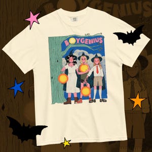 Pode incluir: Camiseta creme com um gráfico de três figuras com chapéus de bruxa segurando lanternas, com o texto "BOYGENIUS" acima delas. A ilustração tem um estilo de história em quadrinhos vintage.