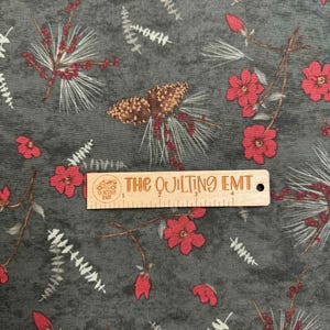 May include: Dark gray fabric with a repeating pattern of red flowers, brown pinecones, and white and gray foliage. A wooden ruler with the text "THE QUILTING EMT" is on the fabric.