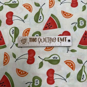 May include: Cream-colored fabric with a repeating pattern of stylized fruits. The design includes red cherries, green pears, orange slices, green apples, and watermelon slices. A wooden ruler with the text "THE QUILTING EMT" is also present.