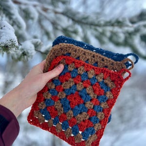 Może przedstawiać: Stos trzech szydełkowych kwadratowych podkładek w odcieniach czerwieni, brązu i niebieskiego. Każda ma pętlę do zawieszenia. Tło to zaśnieżona sceneria zewnętrzna.