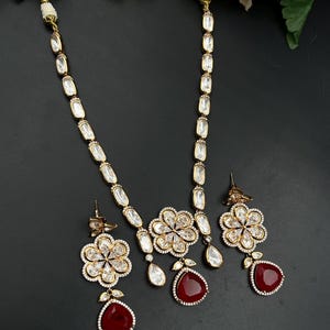 Peut inclure: Un ensemble collier et boucles d'oreilles doré orné de pierres ovales transparentes le long de la chaîne. Le pendentif et les boucles d'oreilles assorties forment des fleurs à six pétales avec des pierres claires et une goutte rouge foncé.