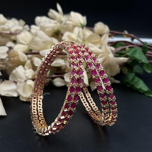 Puede incluir: Dos brazaletes circulares con un marco de metal en tono dorado engastado con filas alternas de piedras rectangulares de color rosa fucsia y pequeñas piedras blancas transparentes. Los brazaletes tienen un diseño geométrico estructurado.