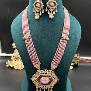 Puede incluir: Un collar de múltiples hilos con pequeñas cuentas rosas y espaciadores dorados, centrado por un gran colgante hexagonal con una piedra rosa central, incrustaciones de color púrpura oscuro y detalles de gotas colgantes, junto a pendientes a juego.