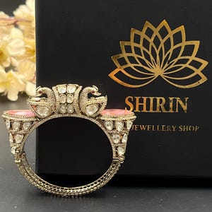 May include: A gold-toned hinged bangle bracelet featuring two swan motifs at the top, accented with clear stones and two oval-shaped pink cabochon gems. The bracelet is displayed in front of a black box with the gold text SHIRIN JEWELLERY SHOP.