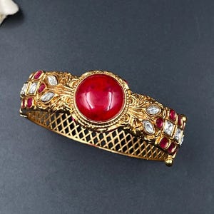 Könnte beinhalten: Ein goldfarbener, aufklappbarer Armreif mit einem großen, runden, glatten roten Cabochon-Stein in der Mitte. Die Metalloberfläche ist mit filigranen, floralen Gravuren versehen und mit kleinen, klaren, marquiseförmigen sowie runden roten Steinen besetzt.
