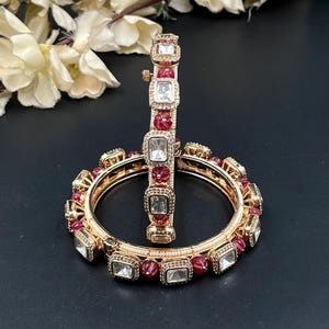 Peut inclure: Deux bracelets rigides dorés ornés d'une alternance de pierres rectangulaires transparentes et de perles rouges texturées. Les bracelets sont agrémentés de petites perles blanches sur le bord intérieur et présentent une conception articulée.