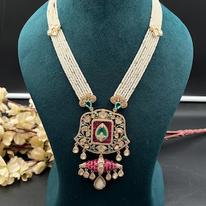 May include: A multi-strand pearl necklace featuring a large, ornate pendant. The pendant is set with a central green stone, surrounded by red stones and clear crystals. A dangling red and gold element hangs from the bottom with small clear drops.