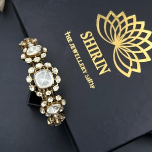 Könnte beinhalten: Ein goldfarbenes Armband mit mehreren klaren, facettierten Steinen in blumenartiger Anordnung. Das Armband liegt auf einer schwarzen Schachtel mit dem goldenen Schriftzug SHIRIN THE JEWELLERY SHOP und einem stilisierten Lotusblüten-Logo.