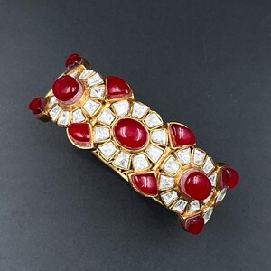 Op de afbeelding: Een goudkleurige armband met een herhalend, bloemachtig patroon. Het ontwerp is bezet met gladde, ovale rode cabochonstenen en heldere, gefacetteerde witte stenen die in een stralend patroon over het gehele oppervlak zijn geplaatst.