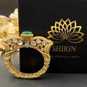 May include: A gold-toned bangle bracelet featuring intricate filigree metalwork, embedded with clear stones, and a prominent rectangular green stone at the top. The bracelet is positioned in front of a black box with a gold lotus logo and the text SHIRIN THE JEWELLERY SHOP.