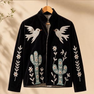 May include: A black velvet jacket featuring a high collar and long sleeves. The front is decorated with white embroidered birds, light blue cacti, and white floral stems. The interior lining is visible and displays a black and white striped pattern.