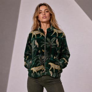 Puede incluir: Una chaqueta de terciopelo verde oscuro con cremallera y un patrón repetitivo de leopardos y palmeras tropicales. La chaqueta tiene cuello alto, mangas largas, una cremallera frontal plateada y puños y dobladillo acanalados, combinada con pantalones vaqueros gris oscuro.