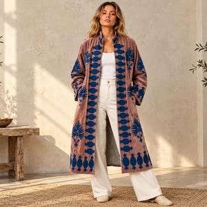 May include: A long, mauve-colored velvet duster coat featuring a prominent dark blue embroidered pattern along the front opening and sleeves. The coat is worn over a plain white tank top and white wide-leg trousers, paired with light-colored shoes.