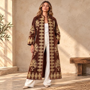 May include: A long, brown velvet duster coat featuring gold-toned embroidery along the front opening and sleeves. The embroidery consists of repeating circular motifs and stylized floral patterns. The coat is worn over a white top and white trousers.