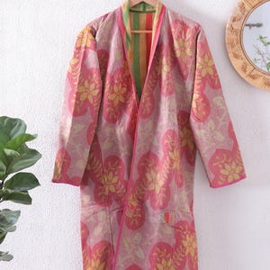 Puede incluir: Una bata larga y abierta con un estampado floral en tonos rosa y amarillo. La bata está colgada de una percha de madera contra una pared blanca. Los bordes de la bata están ribeteados en rosa y el forro interior es a rayas.