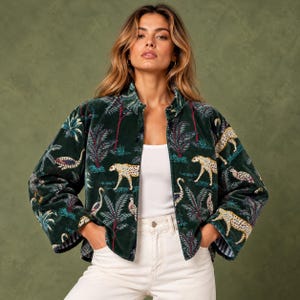 Puede incluir: Una chaqueta de terciopelo verde oscuro con un patrón repetitivo de guepardos, aves y palmeras tropicales. La chaqueta tiene cuello, mangas largas y cierre frontal de botones, combinada con una camiseta blanca y pantalones vaqueros claros.