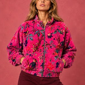 May include: A vibrant hot pink velvet bomber jacket featuring a colorful botanical and bird print. The jacket has a high collar, a full-length front zipper, long sleeves with elasticized cuffs, and a gathered elastic waistband with two side pockets.