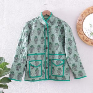 May include: A light green quilted jacket with a floral pattern in shades of green and grey. The jacket features a stand-up collar, four brown buttons, and two front pockets. The edges are trimmed with bright green piping.