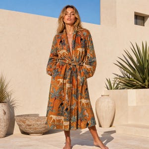 May include: A long-sleeved, calf-length robe in a warm ochre color featuring a repeating pattern of leopards, toucans, and palm trees. The robe has a shawl collar, a tie-waist belt, and a relaxed fit, crafted from a soft, textured fabric.