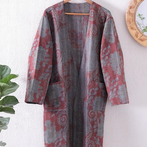 May include: A long, open-front robe with a floral pattern in shades of red and grey. The robe has two front pockets and is hanging on a wooden hanger. The garment appears to be made of a soft, textured fabric.