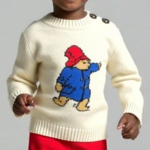 Puede incluir: Un niño lleva un suéter color crema con un diseño de Paddington Bear, combinado con pantalones cortos rojos. El suéter tiene tres botones en el hombro. El diseño de Paddington Bear presenta un sombrero rojo y un abrigo azul. Los pantalones cortos tienen un pequeño diseño de Paddington Bear.
