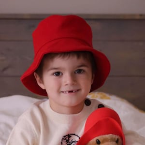 May include: A young child wearing a red bucket hat sits on a white bed and holds a red and blue plush teddy bear. The teddy bear is wearing a red hat and a blue jacket.