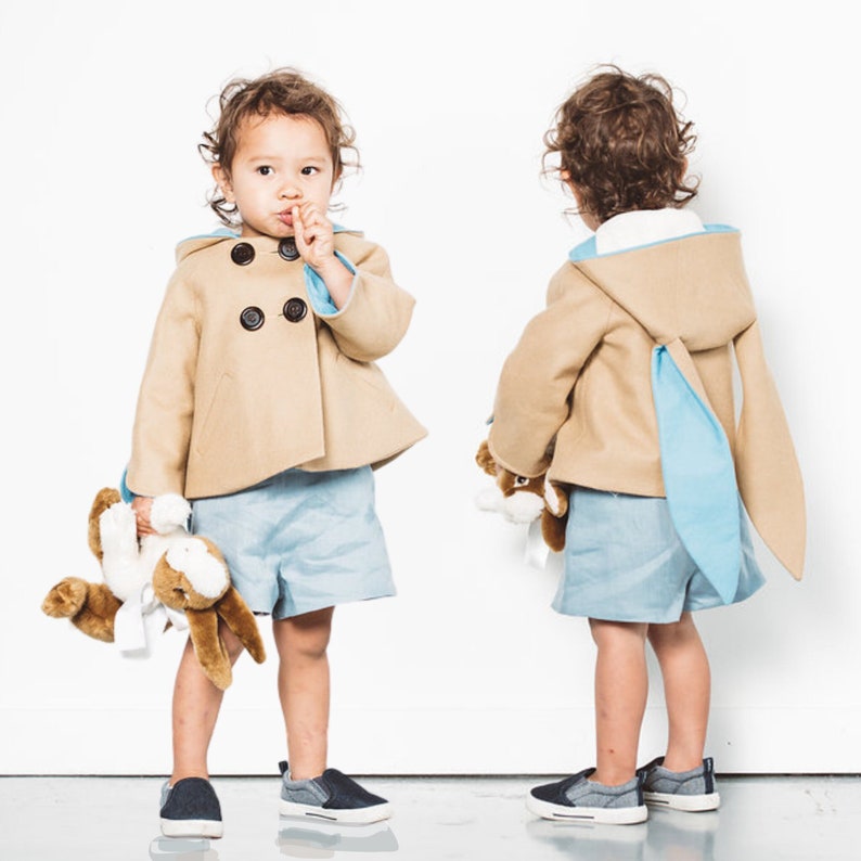 Two toddler boys wearing camel colored wool bunny coats with blue trim.One boy faces front, the other faces away. Each holds a stuffed toy bunny.