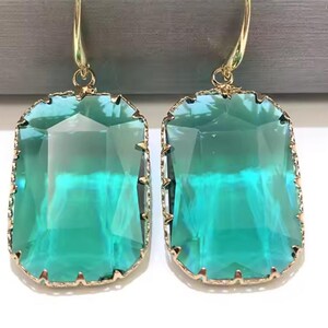 May include: A pair of rectangular, teal-colored earrings with gold-colored trim and hooks. The earrings have a faceted, gem-like appearance, reflecting light. The gold trim has a textured, irregular edge.
