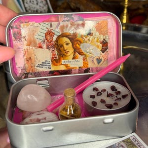 Puede incluir: Una lata de metal plateado que contiene una vela rosa, un pequeño frasco de vidrio con hierbas secas, una piedra de cuarzo rosa lisa y una concha marina. El interior de la tapa muestra un collage con Afrodita y el texto In a dream Aphrodite spoke to me.