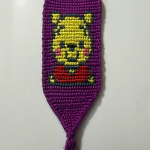 May include: A handmade purple keychain featuring a yellow Winnie the Pooh design with a red shirt. The keychain is made of woven threads and includes a metal keyring.