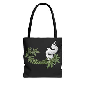 May include: Black tote bag with a black handle. The bag features a green cannabis leaf design with white smoke and the word "Trailwil" in green script. The bag is made of a durable material.