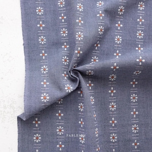 May include: Close-up of a blue fabric with a floral pattern. The fabric features vertical stripes of white and orange flowers and cross-shaped accents. The word "FABLEISM" is printed in white on the fabric.