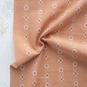 May include: Close-up of peach fabric with a repeating floral pattern. White and brown flowers are arranged in vertical stripes. The fabric is slightly crumpled, showing its texture. The word "FABLEISM" is printed on the fabric.