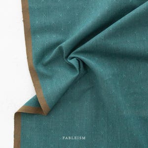 May include: Teal fabric with a subtle cross pattern and a brown border. The fabric is draped, showing the texture and design. The word "FABLEISM" is printed on the fabric.