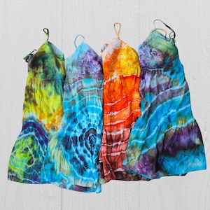 Może przedstawiać: Cztery sukienki tie-dye w żywych kolorach. Sukienki mają różne wzory, w tym wiry i paski, z odcieniami niebieskiego, zielonego, żółtego, pomarańczowego i fioletowego. Każda sukienka ma top w stylu halter i zwiewną spódnicę.