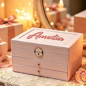May include: A light pink jewelry box with a quilted top lid featuring the name Amellia in raised pink script. The rectangular box has a gold-toned metal lock and two drawers with small gold knobs, resting on a white lace doily near pink roses.