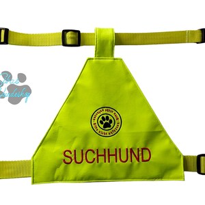 Könnte beinhalten: Eine neongelbe Hunde-Weste mit schwarzen, verstellbaren Riemen und Schnallen. Die Weste hat eine dreieckige Form mit dem rot gestickten Wort "SUCHHUND". Ein rundes Emblem mit einem Pfotenabdruck ist ebenfalls sichtbar.