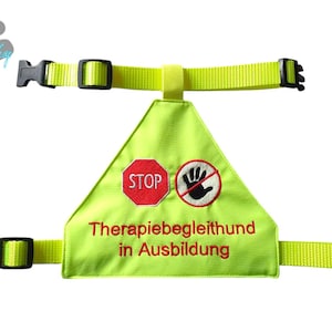 Könnte beinhalten: Eine neongelbe Hunde-Weste mit schwarzen Schnallen und verstellbaren Riemen. Die Weste hat einen dreieckigen Aufnäher mit einem roten "STOP"-Schild und einem Hand-Symbol mit Durchstreichung. Der Text "Therapiebegleithund in Ausbildung" ist rot gestickt.