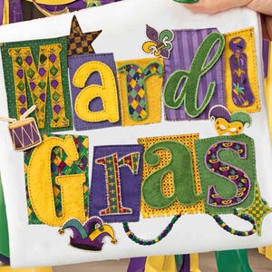 May include: A white fabric item with the words "Mardi Gras" in colorful, appliqued letters. The letters are in yellow and green, with purple and gold accents. Decorative elements include a drum, a star, and a jester's hat.