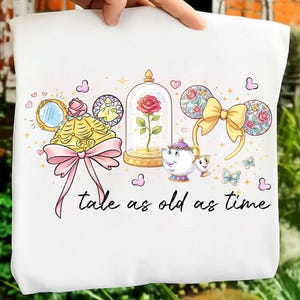 以下が含まれることがあります： 白地のTシャツに、美女と野獣のキャラクターが描かれたカラフルなデザイン。ガラスドームに入ったバラ、ティーカップ、リボン付きのミッキーマウスの耳が特徴です。「tale as old as time」の文字が黒で書かれています。