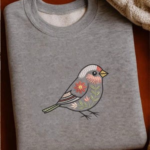 Puede incluir: Una sudadera gris con un dise&ntilde;o de p&aacute;jaro bordado. El p&aacute;jaro est&aacute; adornado con patrones florales en tonos rosa, naranja y verde. El pico del p&aacute;jaro es amarillo y el dise&ntilde;o est&aacute; sobre la tela gris de la sudadera.
