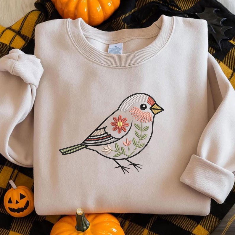 Puede incluir: Sudadera beige con un dise&ntilde;o de p&aacute;jaro bordado. El p&aacute;jaro est&aacute; decorado con patrones florales y de hojas en rojo, verde y blanco. La sudadera tiene mangas largas y cuello redondo.
