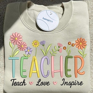 Könnte beinhalten: Beiges Sweatshirt mit dem Wort "TEACHER" in bunten Buchstaben gestickt, jeder Buchstabe mit floralen Designs verziert. Darunter die Worte "Teach Love Inspire". Ein rundes Etikett mit dem Markennamen "banagoya" ist angebracht.