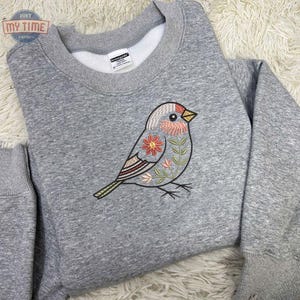 Puede incluir: Sudadera gris con cuello redondo con un dise&ntilde;o de p&aacute;jaro bordado. El p&aacute;jaro tiene la cabeza roja, el pecho rosa y detalles florales en rosa, naranja y verde. La sudadera tiene el texto "DON'T MY TIME" en un logotipo circular.