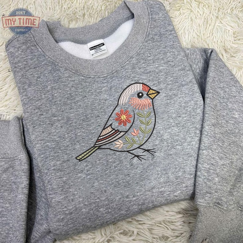 Puede incluir: Sudadera gris con cuello redondo con un dise&ntilde;o de p&aacute;jaro bordado. El p&aacute;jaro tiene la cabeza roja, el pecho rosa y detalles florales en rosa, naranja y verde. La sudadera tiene el texto "DON'T MY TIME" en un logotipo circular.