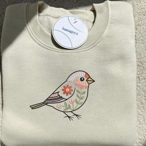 Puede incluir: Sudadera verde claro con cuello redondo y etiqueta redonda con la inscripci&oacute;n "banagoya". La sudadera presenta un dise&ntilde;o de p&aacute;jaro bordado con detalles florales en rosa, rojo y verde.