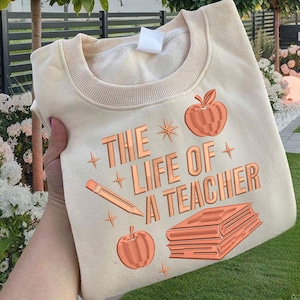 May include: Cream-colored sweatshirt with the text "THE LIFE OF A TEACHER" in a copper-colored, embossed font. The design includes illustrations of an apple, a pencil, and a stack of books. The sweatshirt is folded.