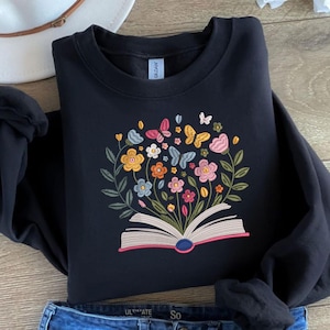 May include: Black sweatshirt featuring an embroidered design of an open book with colorful flowers and butterflies. The design includes yellow, orange, pink, blue, and green elements. The sweatshirt is folded on a light-colored surface.