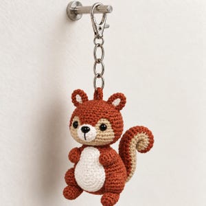 May include: A crochet squirrel keychain featuring a rust-colored body, a cream-colored belly and face, and a bushy tail. The squirrel has black bead eyes and a small black stitched nose. It is attached to a silver-toned metal lobster clasp.