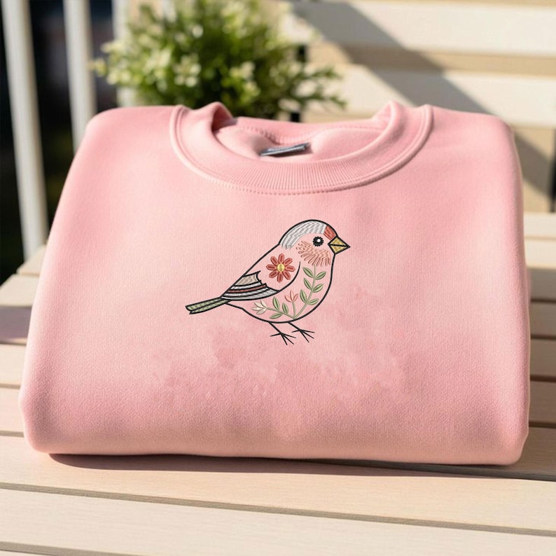 Puede incluir: Una sudadera rosa claro doblada con un dise&ntilde;o de p&aacute;jaro bordado. El p&aacute;jaro tiene un cuerpo rosa, alas grises y un pico amarillo, con detalles florales y de hojas. La sudadera est&aacute; hecha de un material suave y es adecuada para uso casual.